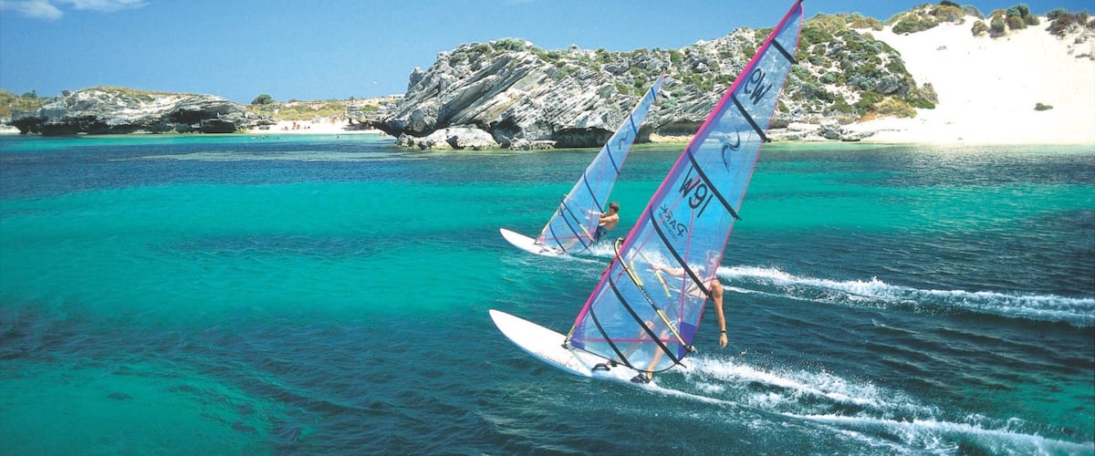 Two people windsurfing on turquoise waters with rocky cliffs in the background.