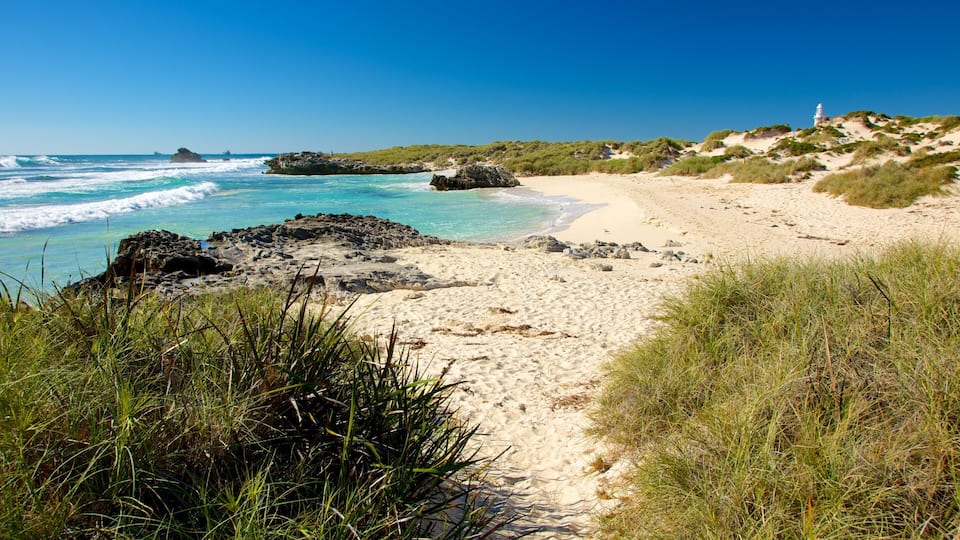 A sandy beach with clear turquoise waters, rocky outcrops, and a lighthouse in the distance.
