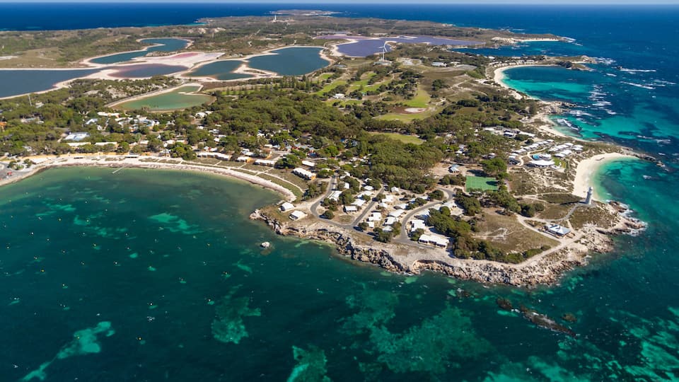 An aerial view of a coastal area with clear turquoise waters, a lighthouse, and a mix of residential and natural landscapes.