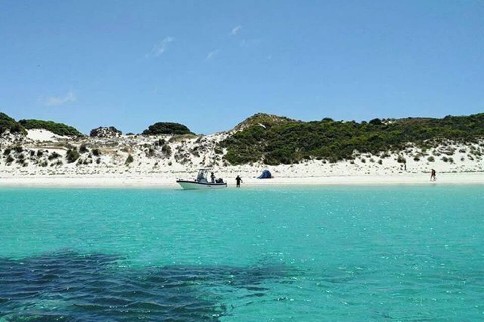 A beach with clear turquoise water, white sand, and green hills in the background.
