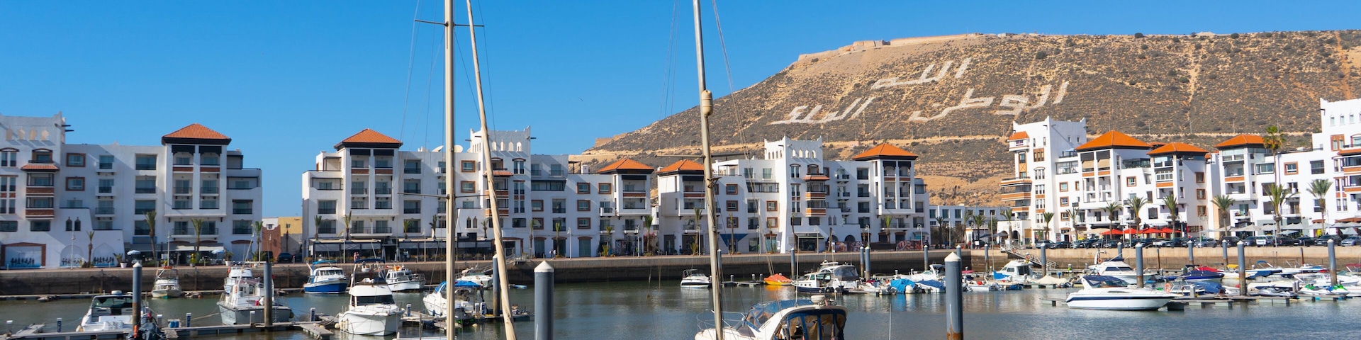A marina with several boats docked, a clear blue sky, and a hilly landscape in the background.