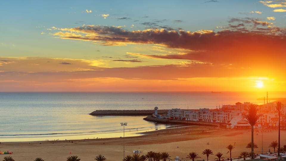 A coastal cityscape at sunset with a beach, buildings, and a distant mountain.