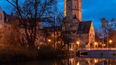A historic tower reflecting in a calm river, surrounded by trees and buildings.