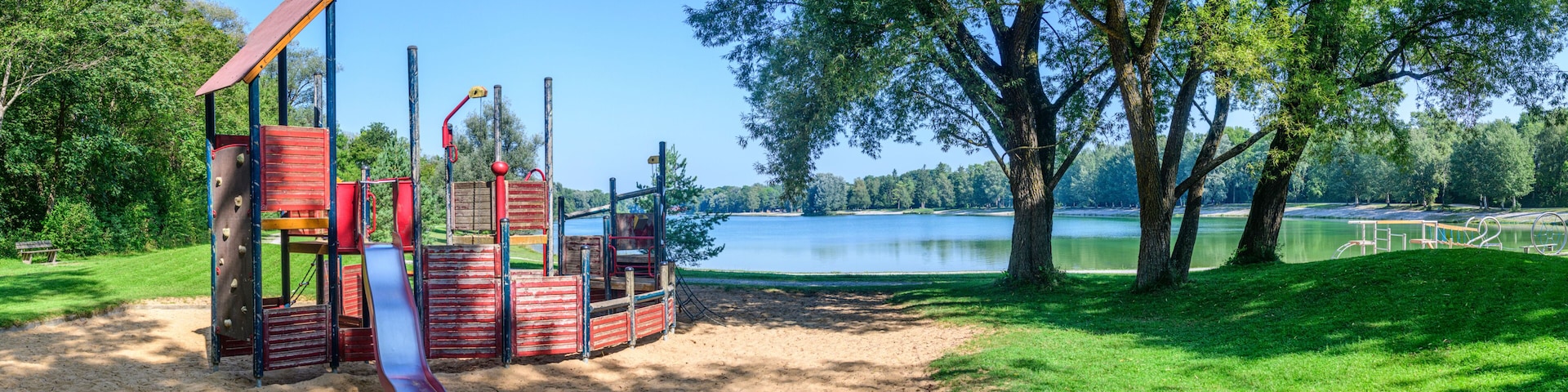 A playground with a slide and climbing structures, a sandy area, and a lake with trees in the background.