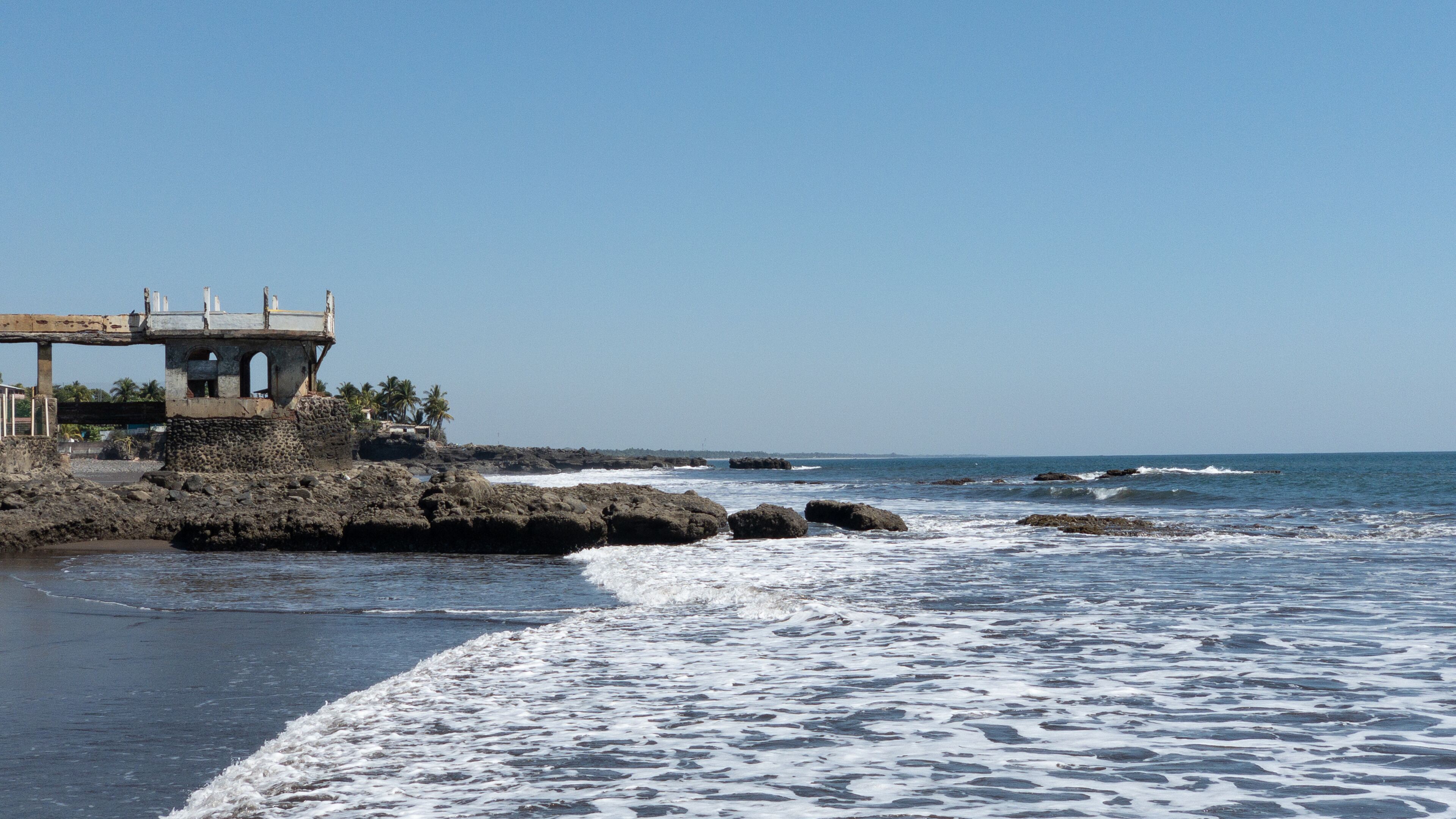 Uma costa rochosa com ondas quebrando na praia, uma construção desgastada com uma plataforma de observação e palmeiras ao fundo.