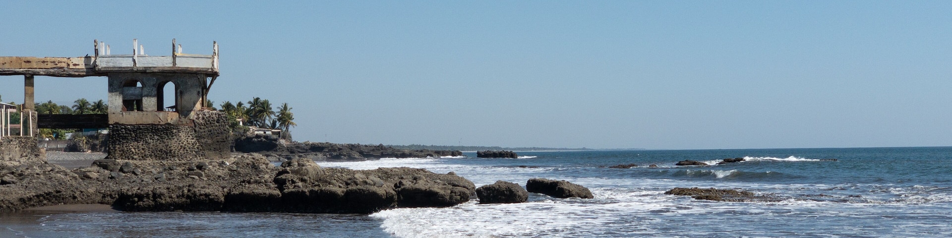 Uma costa rochosa com ondas quebrando na praia, uma construção desgastada com uma plataforma de observação e palmeiras ao fundo.