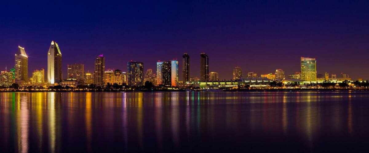 A nighttime city skyline with illuminated buildings reflecting on the water.