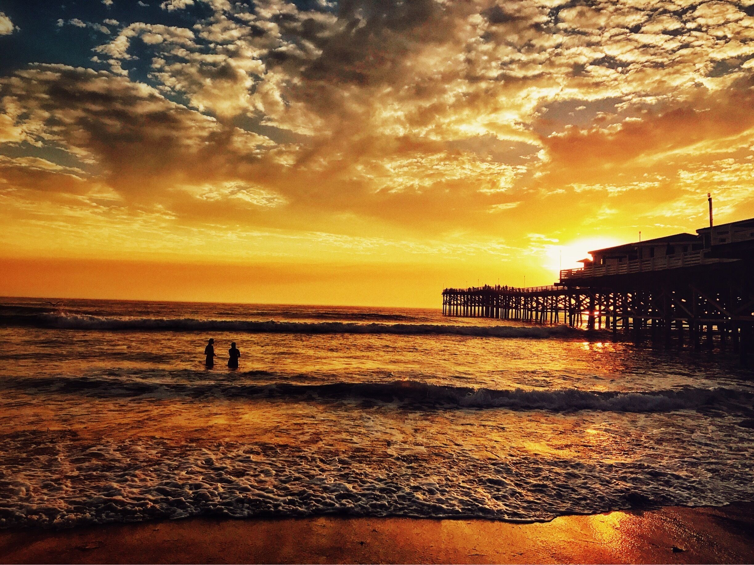 A sunset over a beach with waves and a pier.