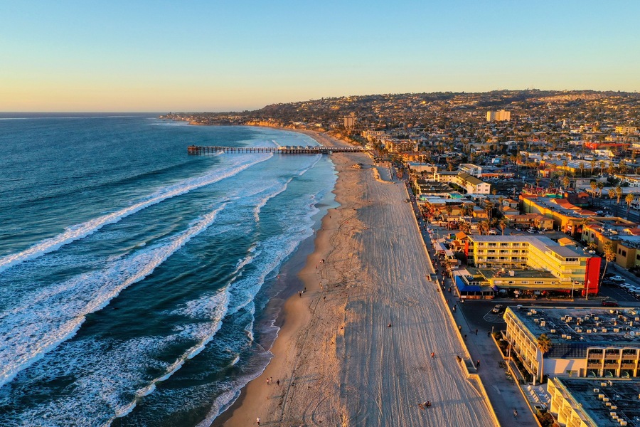 A coastal cityscape with a beach, ocean waves, a pier, and urban buildings.