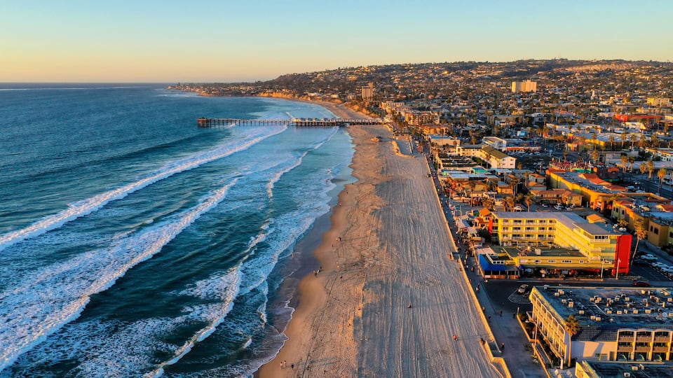A coastal cityscape with a beach, ocean waves, a pier, and urban buildings.