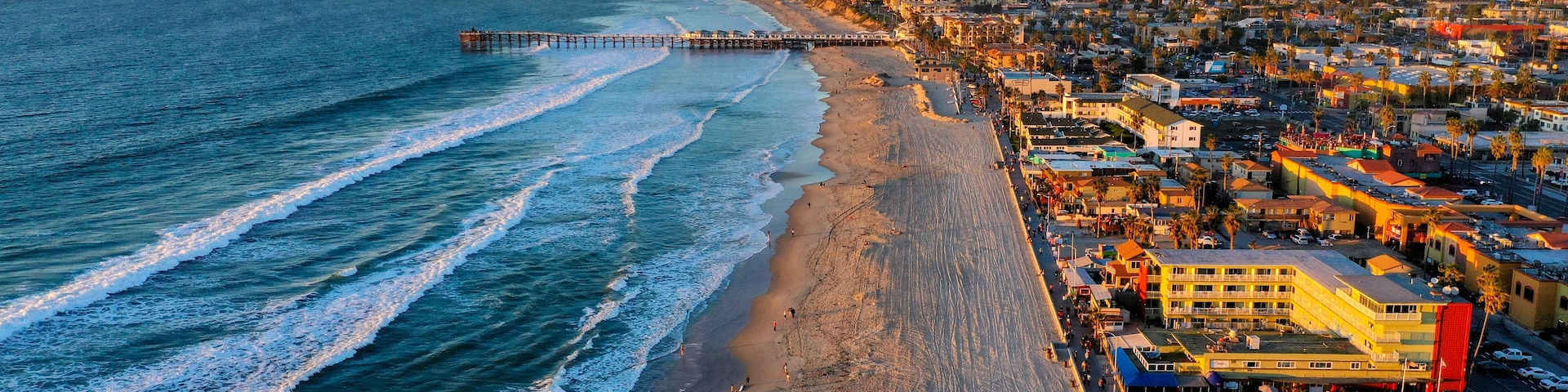A coastal cityscape with a beach, ocean waves, a pier, and urban buildings.