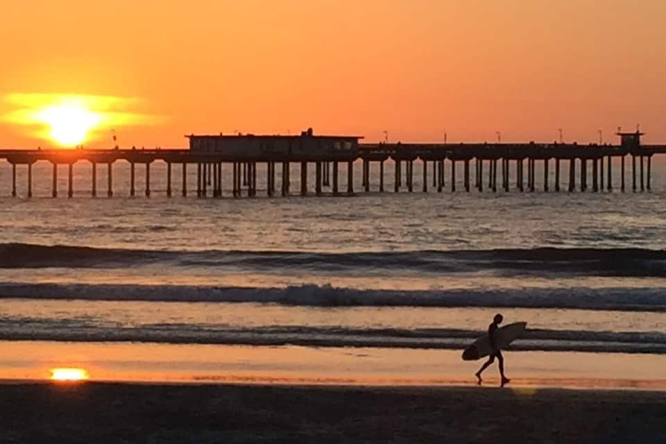 Um pôr do sol sobre um cais que se estende para o oceano, com uma pessoa carregando uma prancha de surf caminhando na praia.