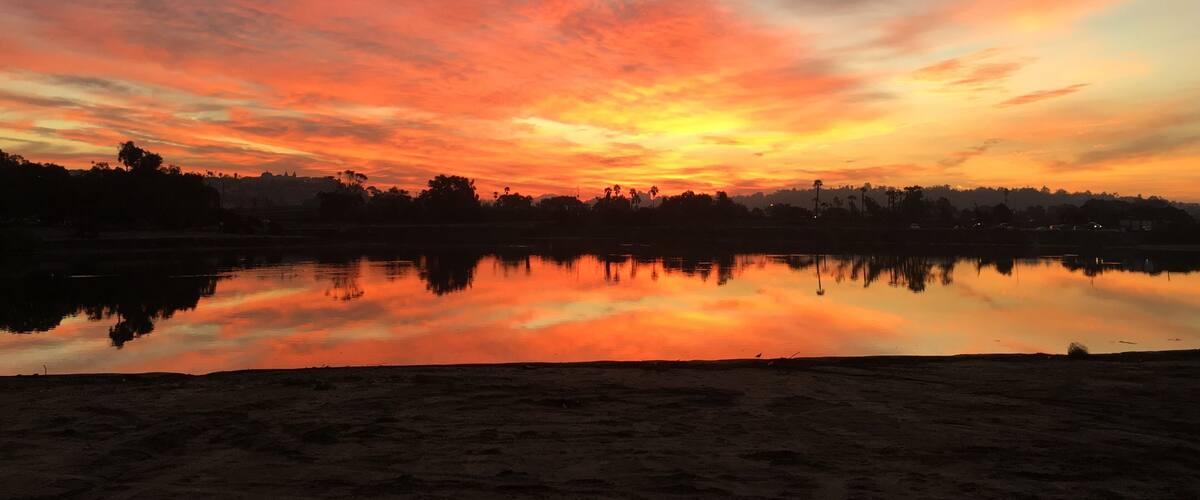 A sunset over a calm lake with a silhouette of trees and buildings on the horizon.