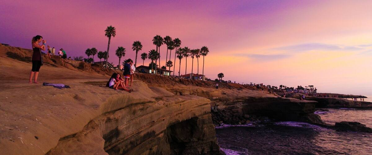 A rocky coastline with palm trees and people at sunset.