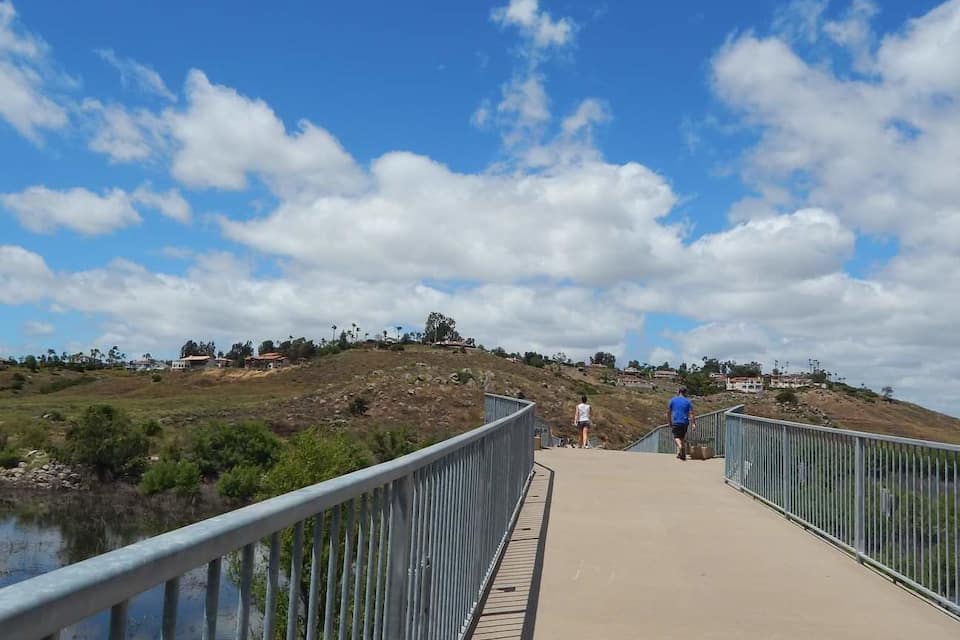 A pedestrian bridge over a body of water with a metal railing, surrounded by greenery and a hilly landscape.