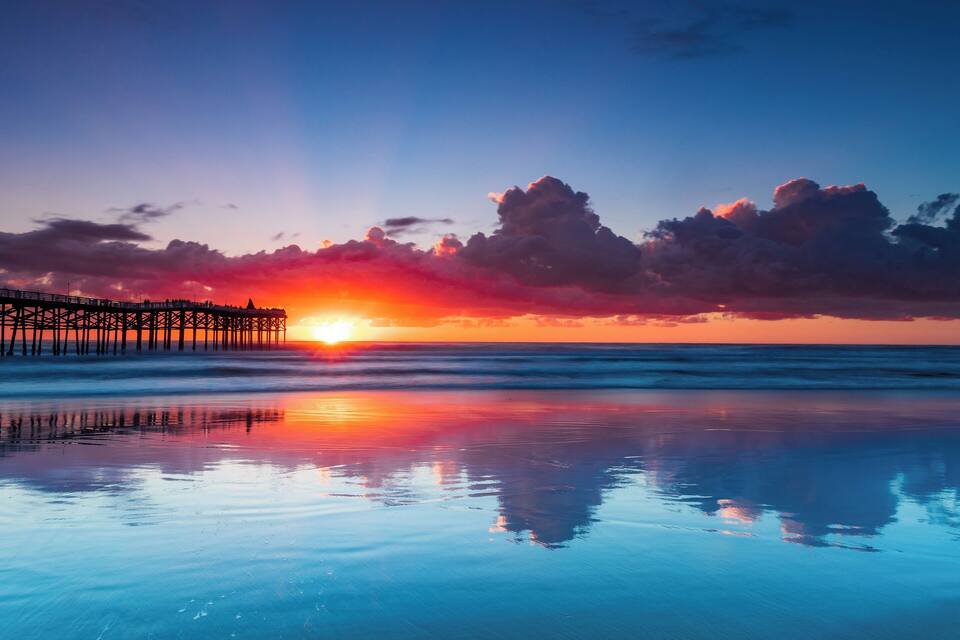 Un atardecer sobre una playa con un muelle que refleja el cielo.