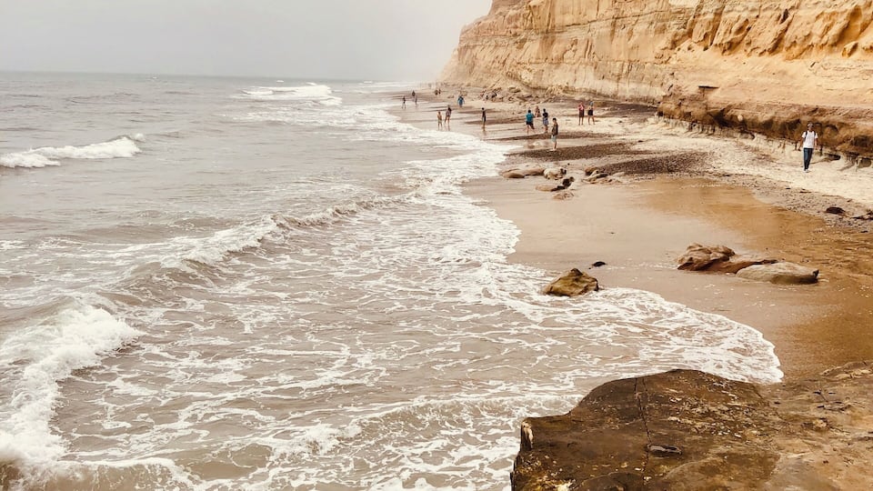 Uma praia rochosa com ondas quebrando na costa e pessoas caminhando pela orla.