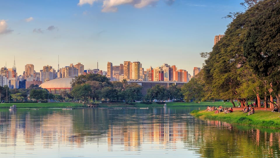 A city skyline with a large stadium, a lake reflecting buildings, and trees along the shore.