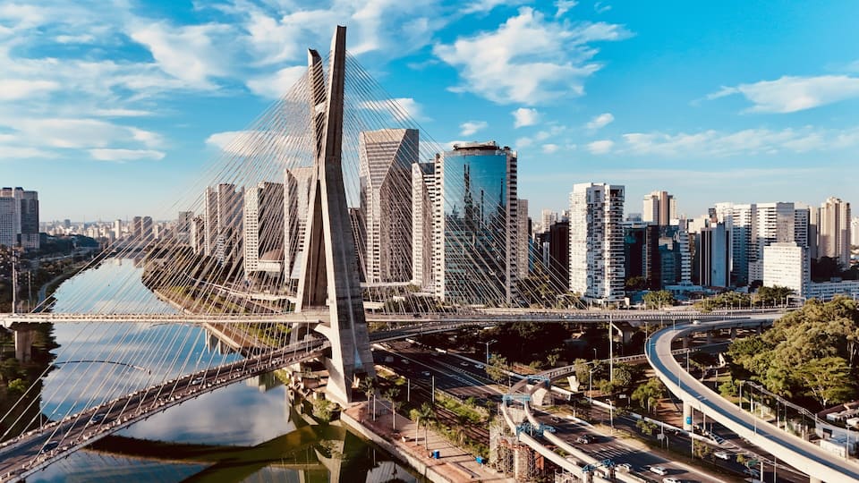 A modern cityscape with a prominent cable-stayed bridge over a river, surrounded by high-rise buildings and a complex network of roads and overpasses.
