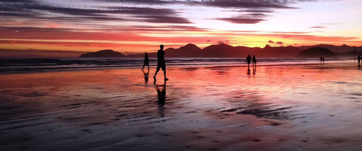 Una playa al atardecer, con siluetas de personas caminando, superficie de arena húmeda y montañas distantes.