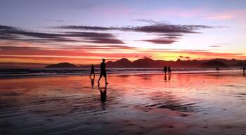 Una playa al atardecer, con siluetas de personas caminando, superficie de arena húmeda y montañas distantes.