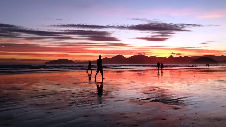 A beach at sunset with silhouettes of people walking, a wet sand surface, and distant mountains.