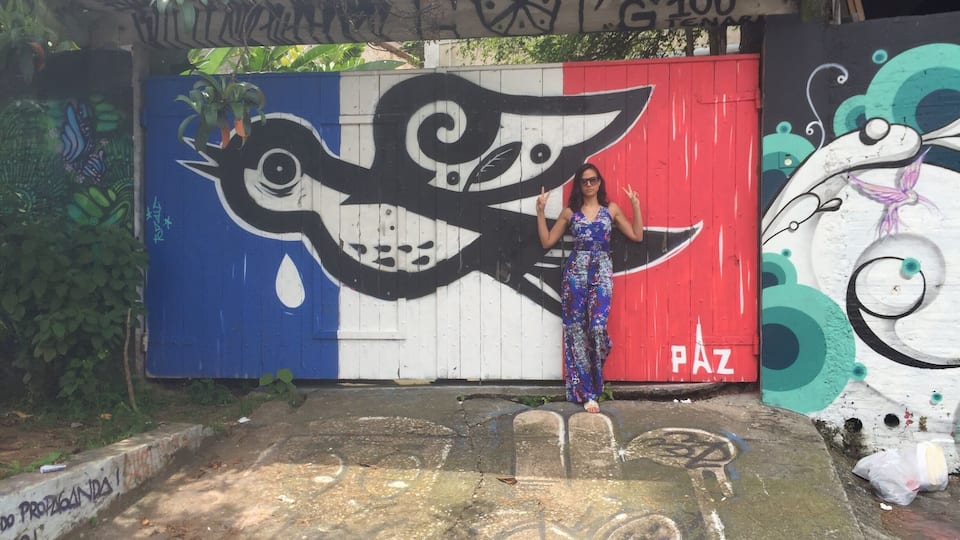 A person standing in front of a mural with a large eye design, a red and white section with the word "PAZ", and graffiti on a concrete wall.