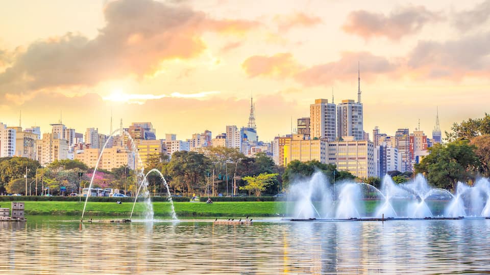 A cityscape with a park featuring fountains and a body of water reflecting the skyline.