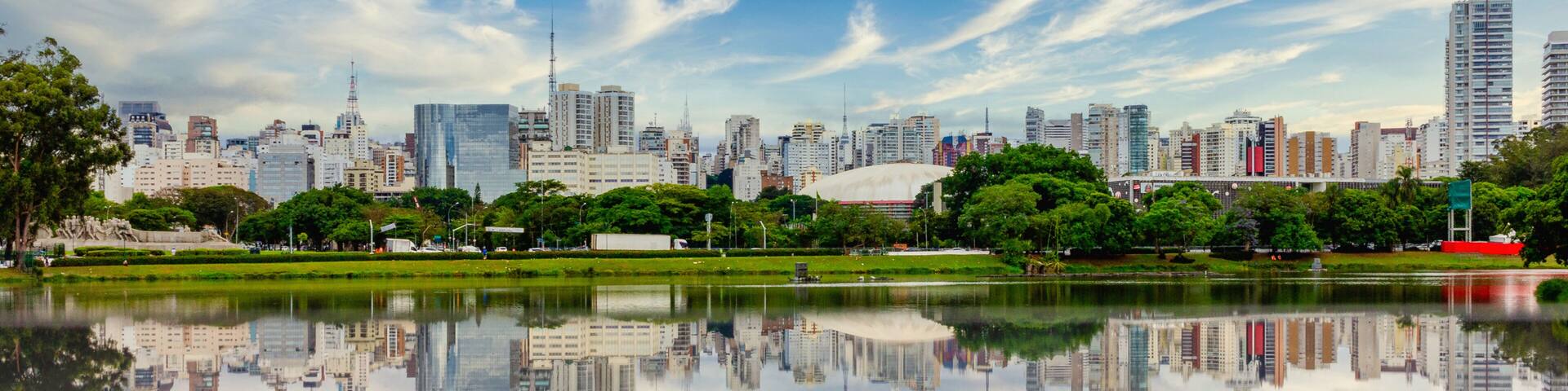 A city skyline reflected in a calm body of water, surrounded by greenery and a clear sky.