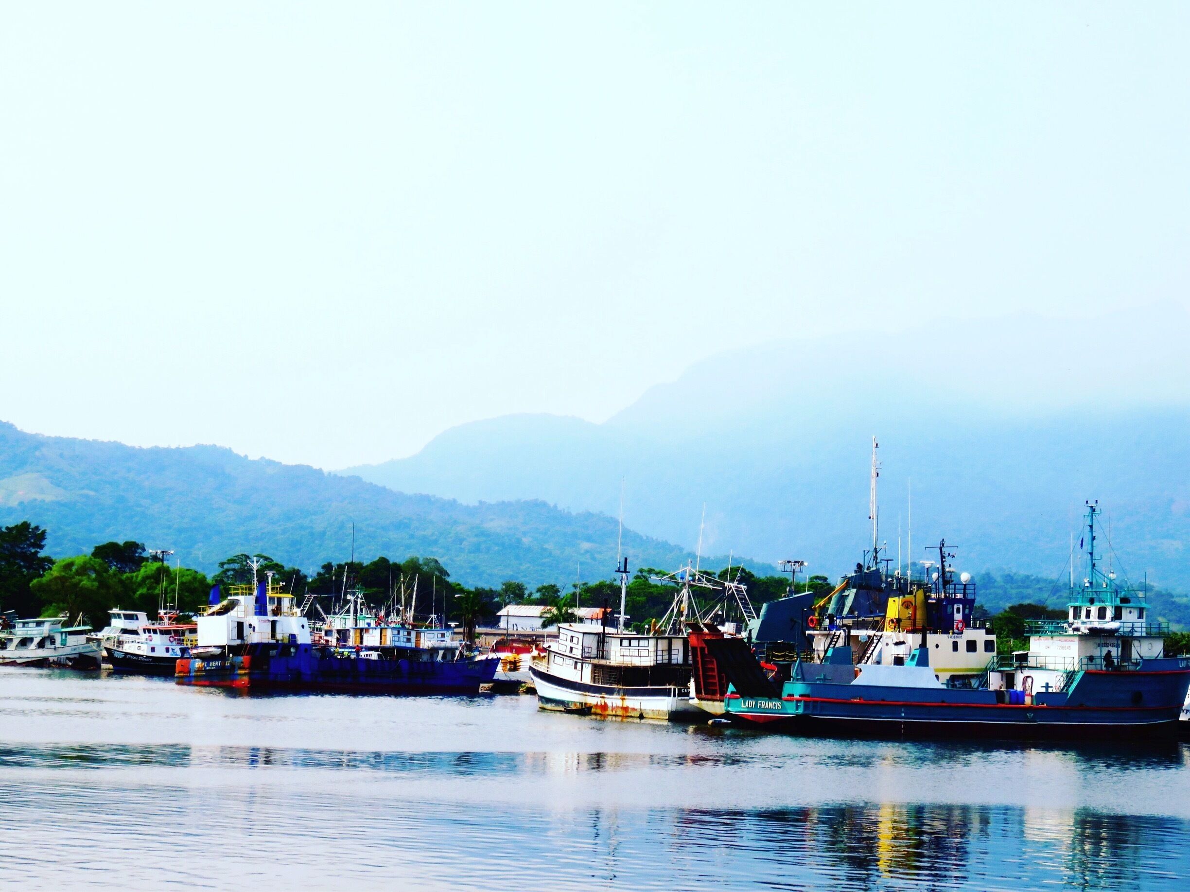 A harbor with several fishing boats docked, a lighthouse, and a mountainous backdrop.