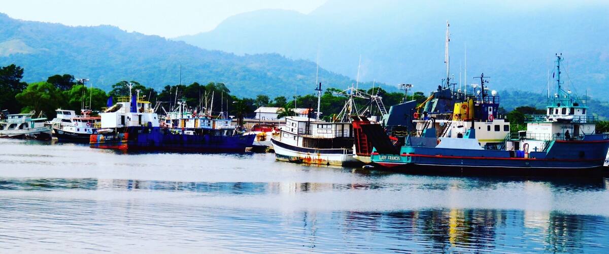 A harbor with several fishing boats docked, a lighthouse, and a mountainous backdrop.