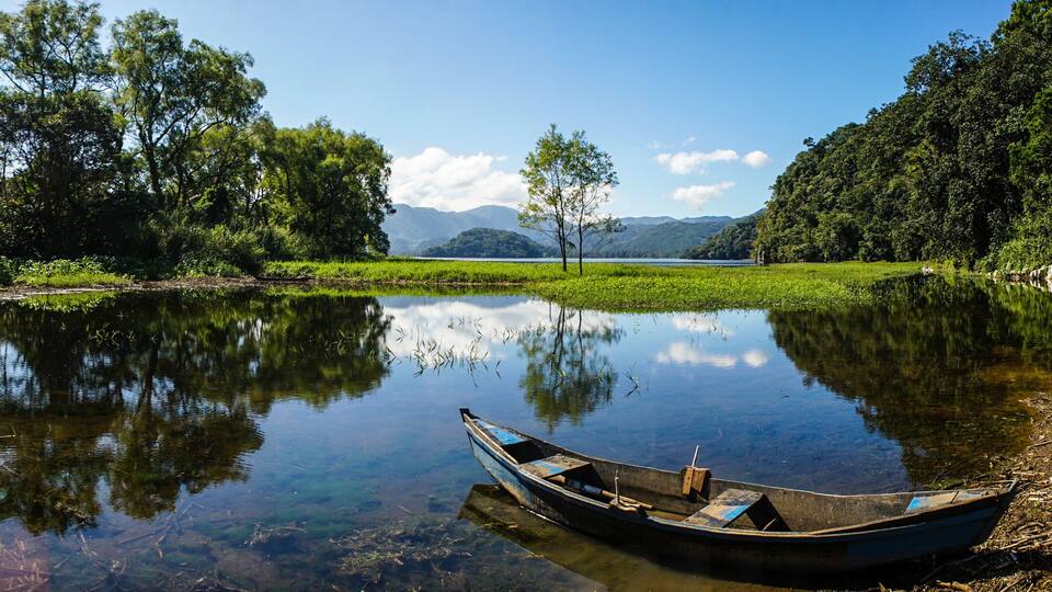 A calm river with a small boat, surrounded by lush greenery and distant mountains.