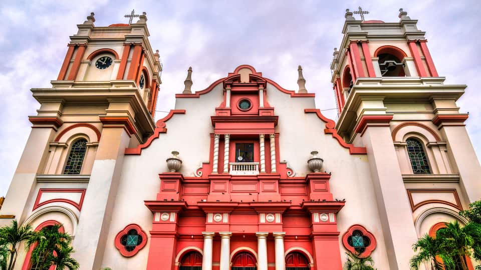 A church with two bell towers, red and white facade, and palm trees in front.