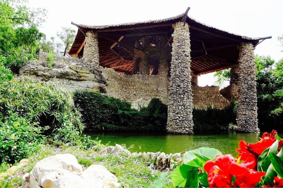 Un puente de piedra con techo de paja cruzando un río, rodeado de abundante vegetación y plantas en flor.
