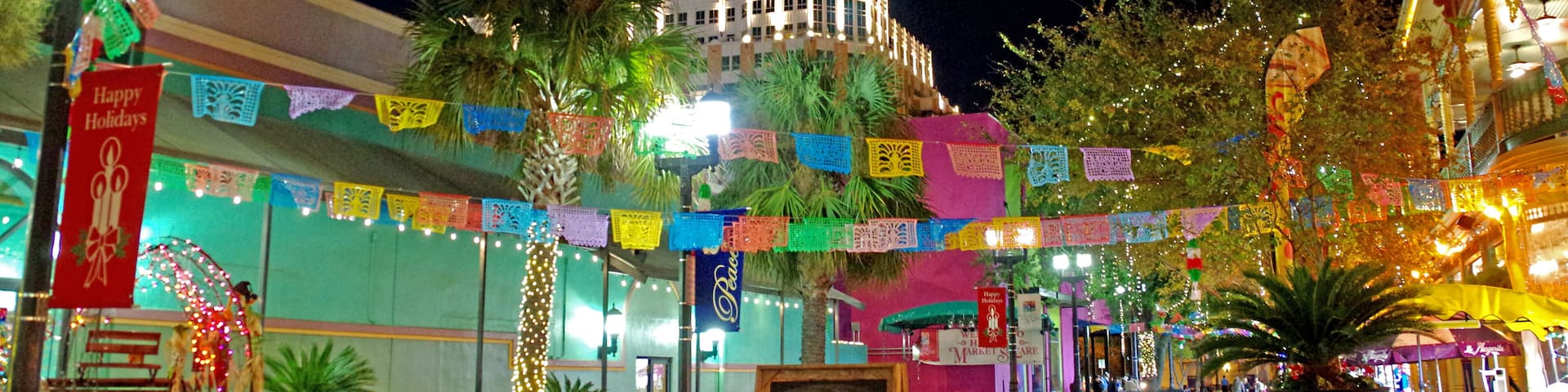 A brightly lit street with colorful flags, a building with a tower, and signs for restaurants.