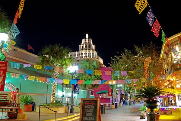 Uma rua bem iluminada com bandeiras coloridas, um edifício com torre e placas indicando restaurantes.