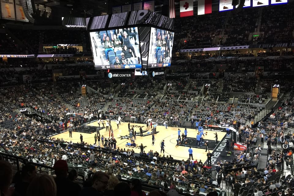 A basketball arena with a large screen displaying a game, a basketball court, and a crowd of spectators.