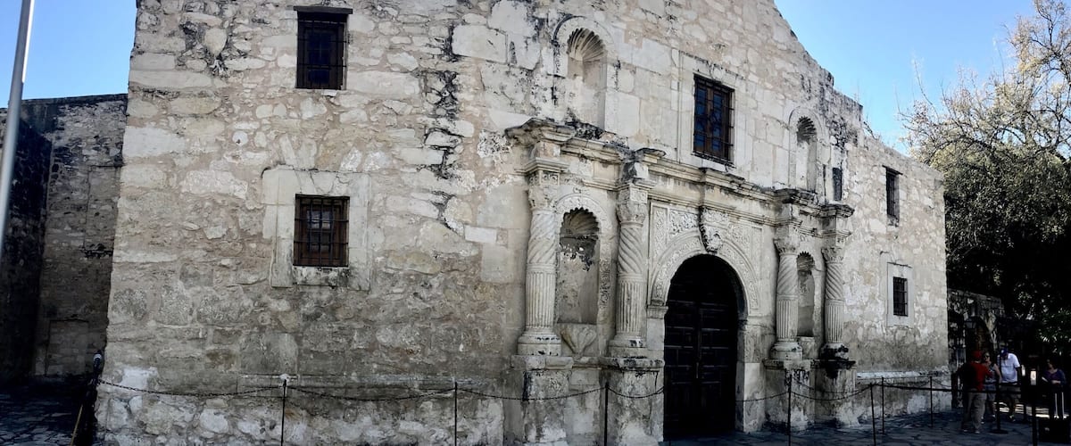 A historic stone building with arched windows and a large door, surrounded by a cobblestone plaza and a flagpole.