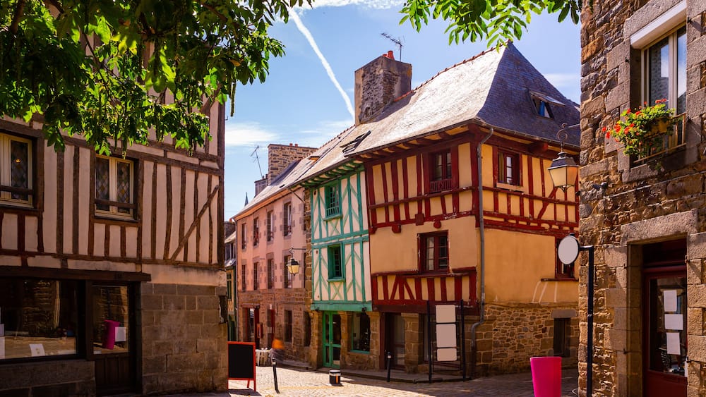 Une rue pavée bordée de bâtiments aux façades à colombages aux couleurs vives, un lampadaire et une poubelle rouge.