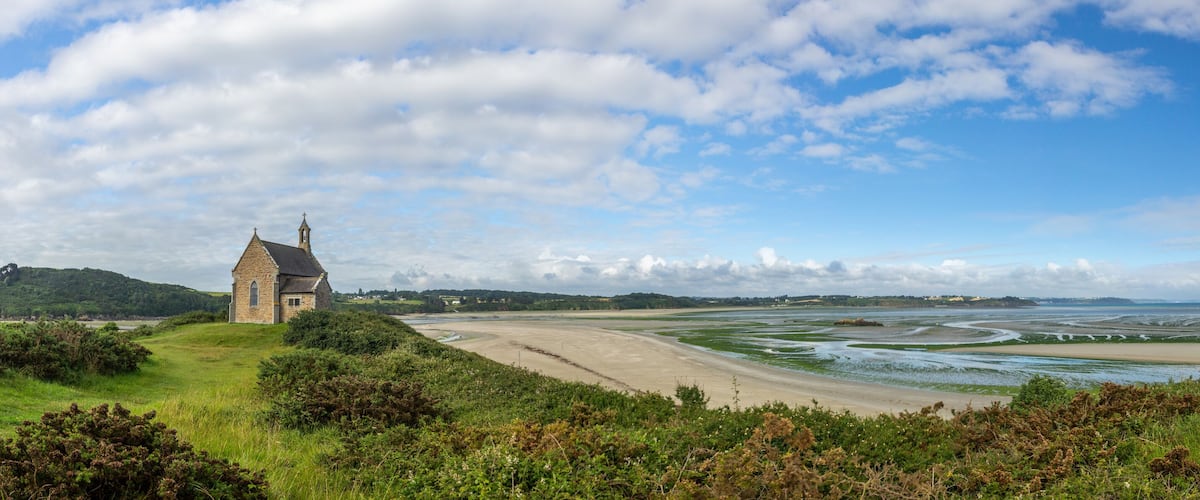 A coastal landscape with a stone church, green hills, and a wide sandy beach with tidal flats.