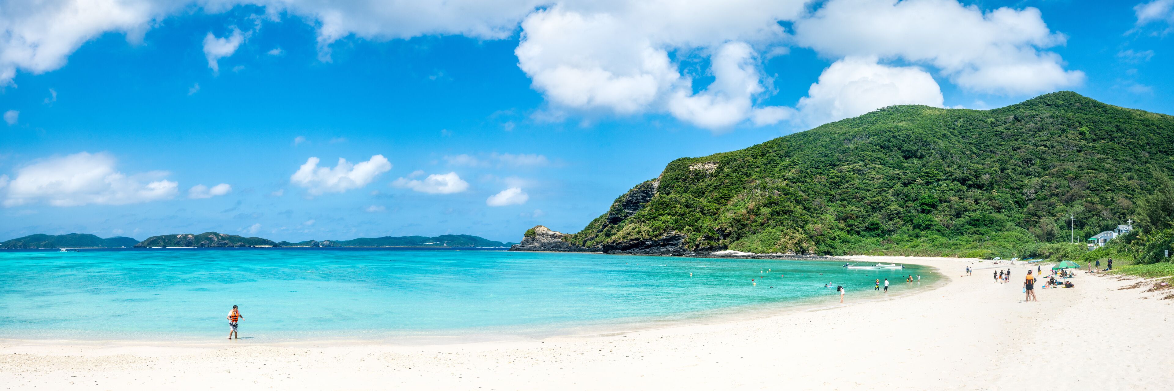 A beach with clear turquoise water, a sandy shore, and a green hill in the background.