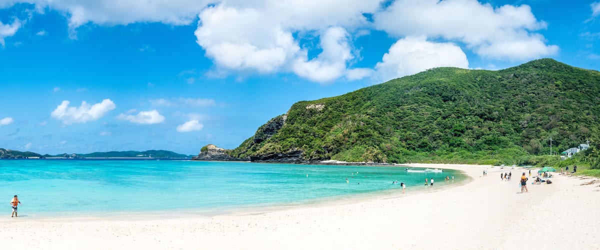 A beach with clear turquoise water, a sandy shore, and a green hill in the background.