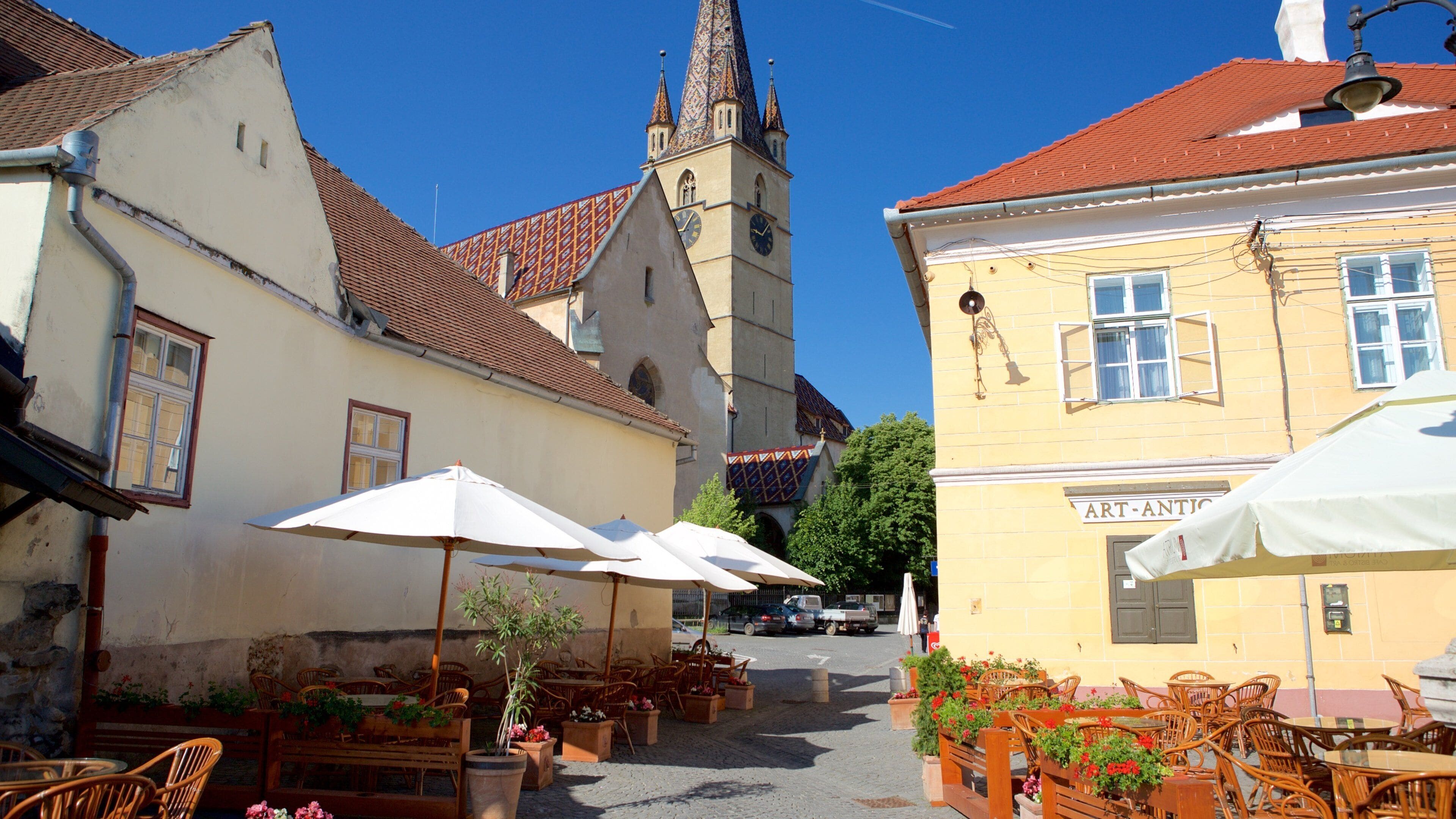 A narrow street with buildings, a church tower, outdoor seating with umbrellas, and a sign reading "ART-ANTIC".