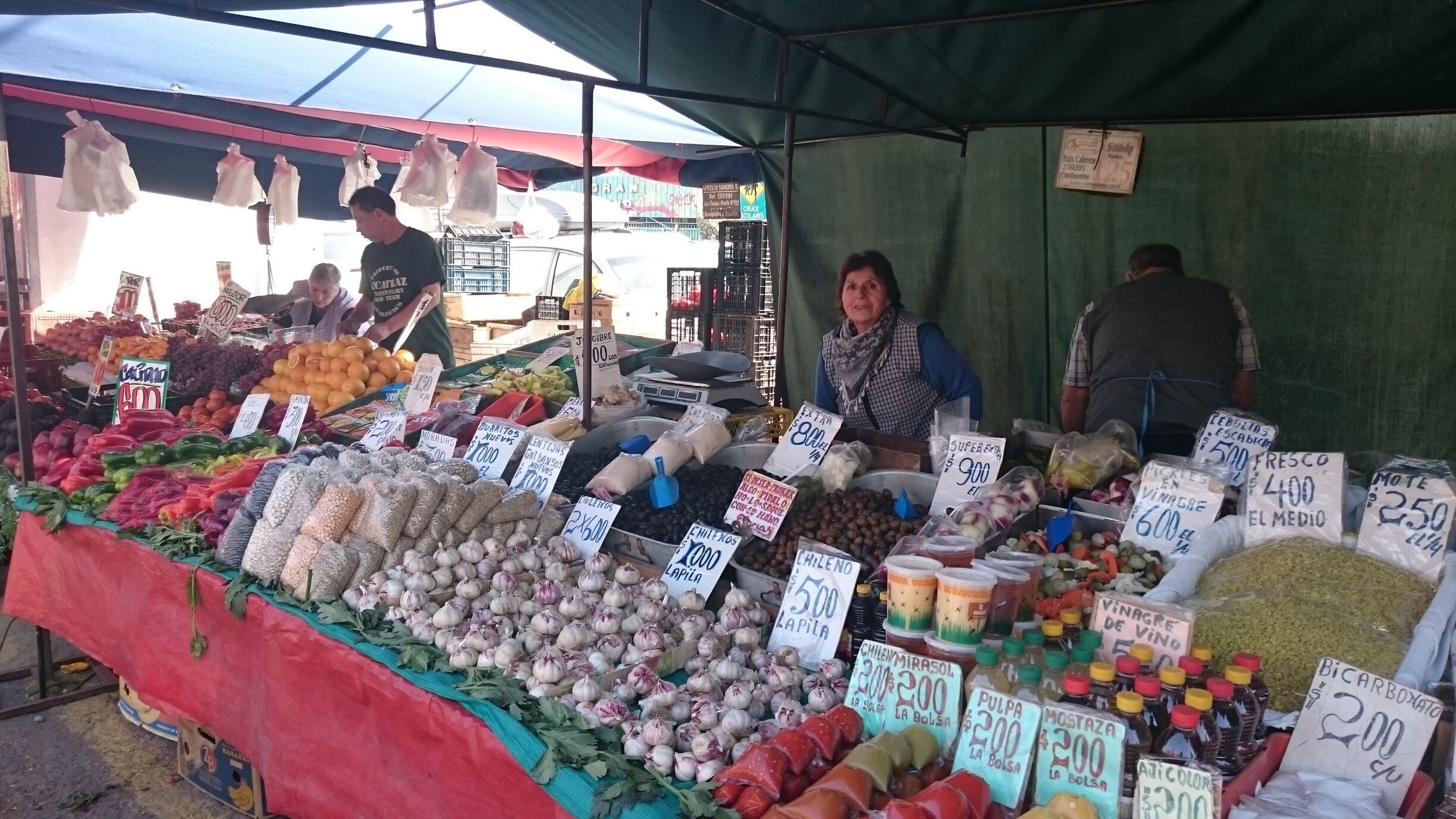 A market stall with various vegetables and fruits, including onions, garlic, and tomatoes, with price tags visible.