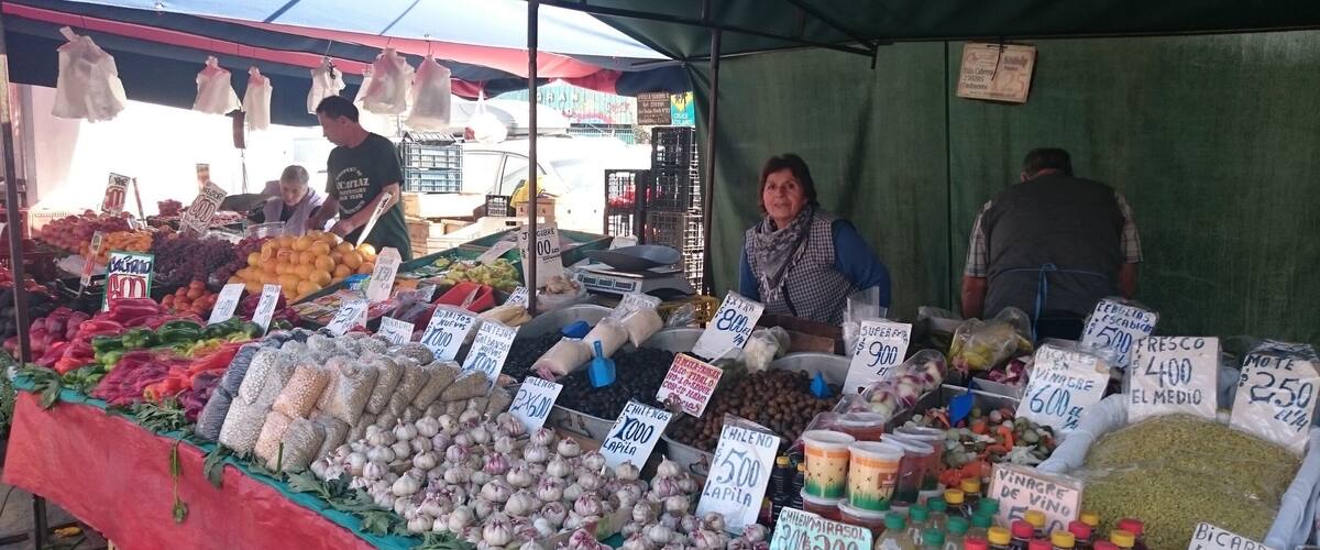 A market stall with various vegetables and fruits, including onions, garlic, and tomatoes, with price tags visible.