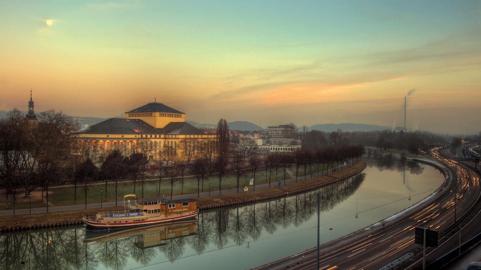 A river with a boat, a bridge, and a cityscape with buildings and a tower.