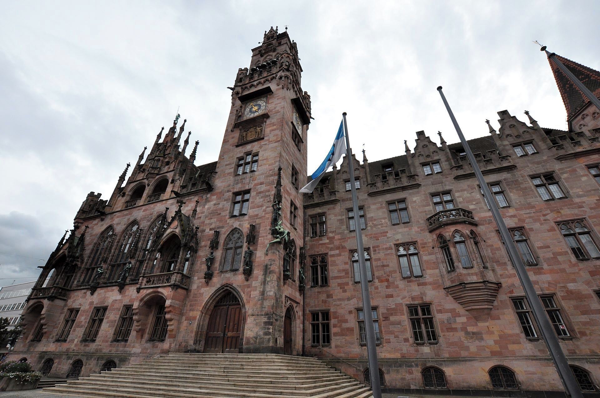 A historic building with intricate Gothic architecture, a prominent clock tower, and a flagpole with a flag.