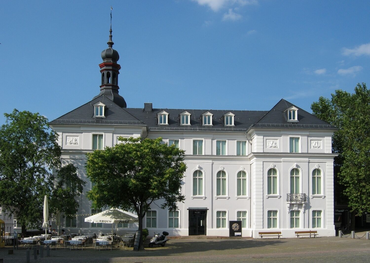 A large white building with a tower, multiple windows, and outdoor seating.