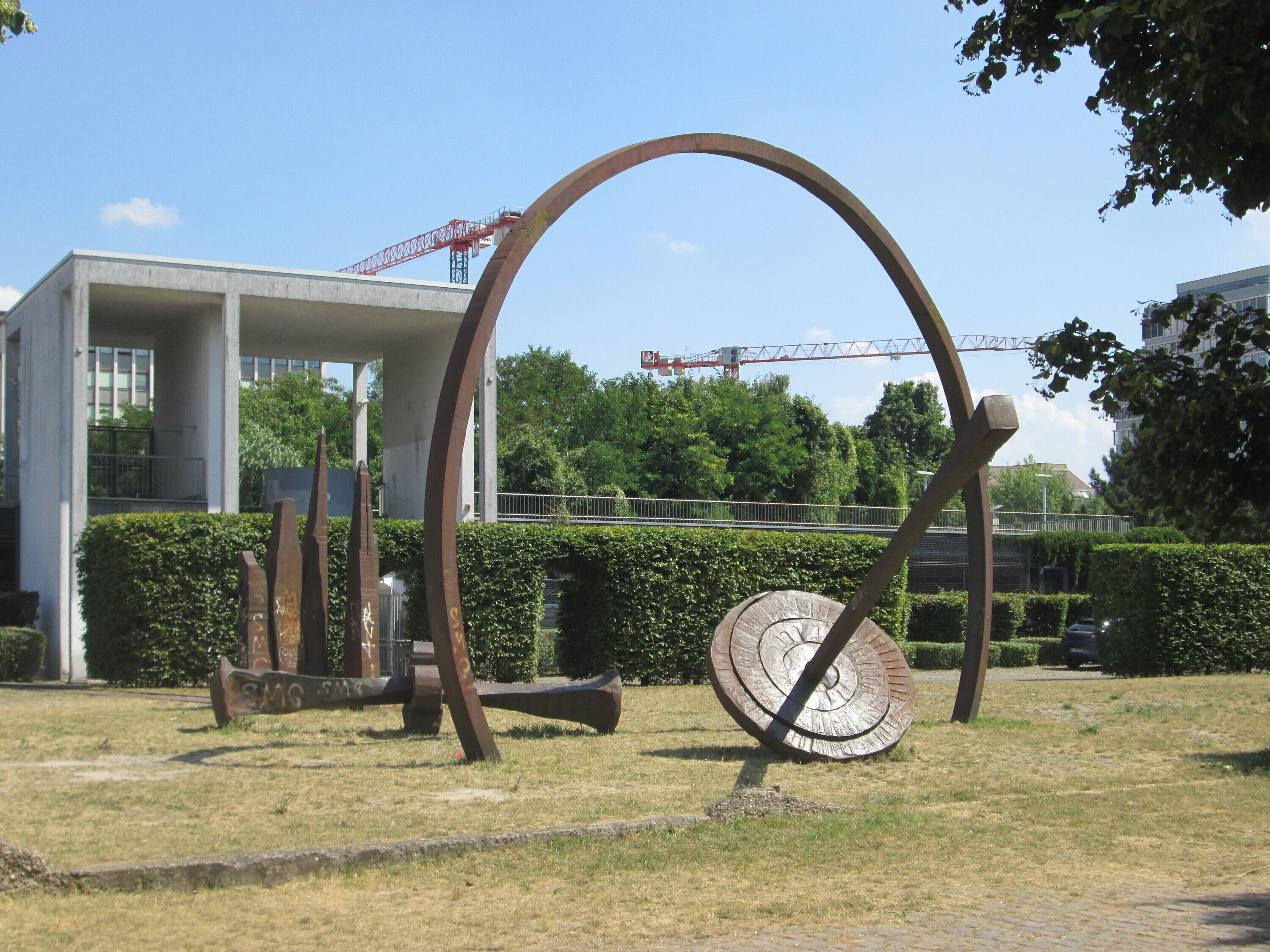 A sculpture with a circular element and a large arch in a grassy area.
