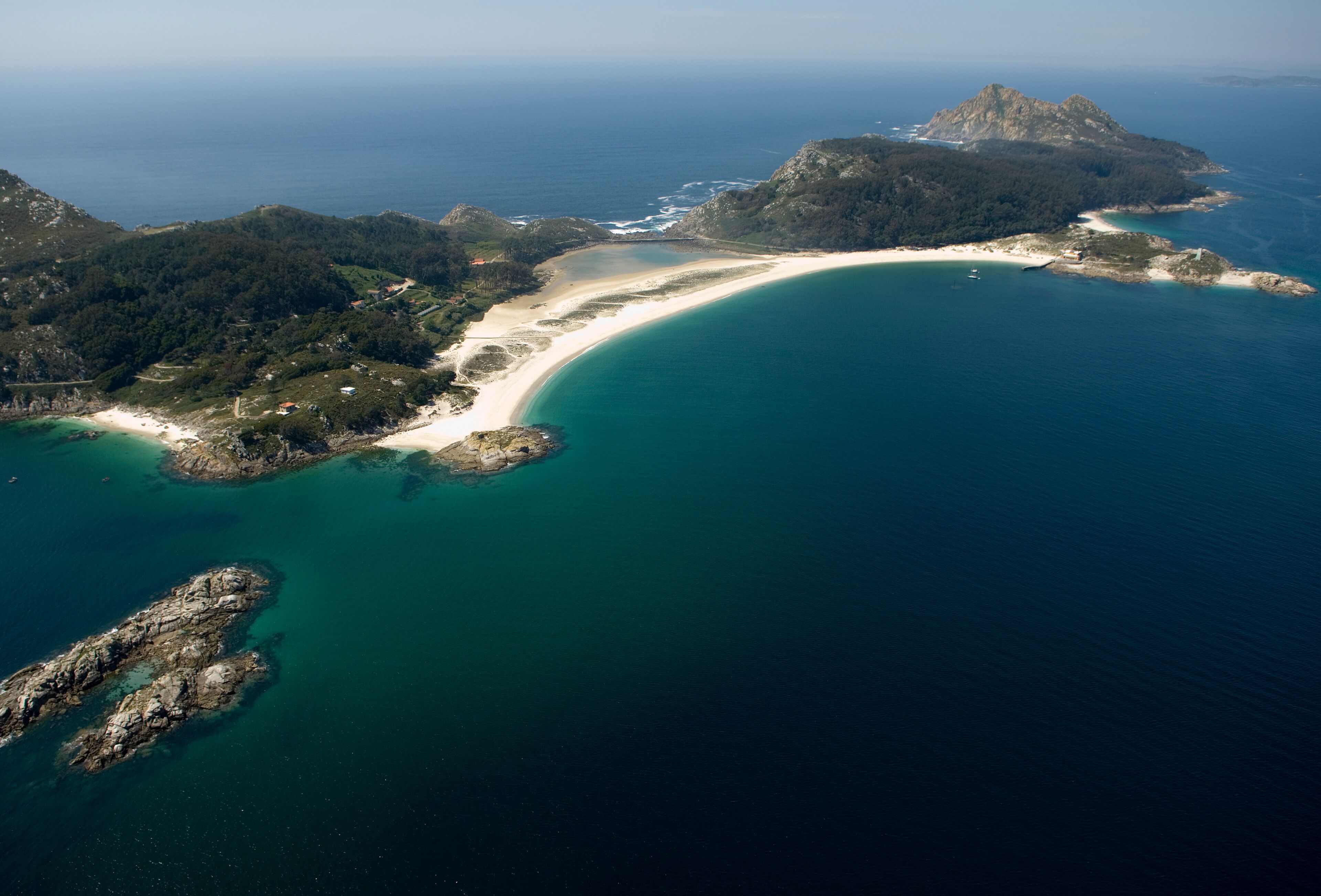 Vista aérea de un paisaje costero con una playa de arena, aguas turquesas cristalinas y un promontorio rocoso destacado.